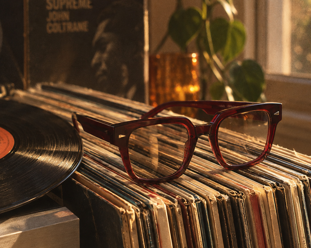 Rustic brown acetate square-frame eyeglasses resting on a stack of vinyl records next to a turntable, with a record sleeve and plant in the background.